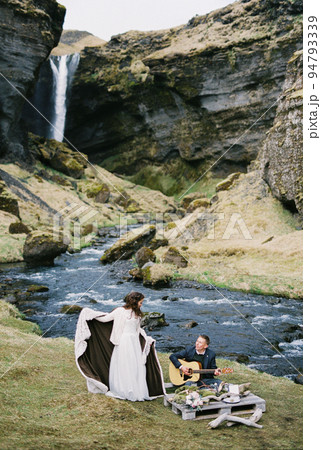 Groom plays the guitar next to the bride standing in a blanket on the river bank 94793339