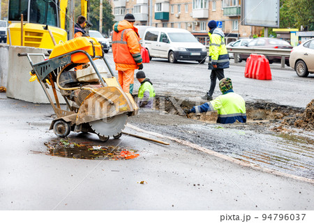 A yellow power cutter with a diamond blade against the backdrop of a road crew working to repair storm drains on an autumn day. 94796037
