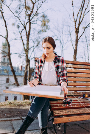 Young woman artist draws a picture while sitting on a bench. Painter artist holds a wooden tablet for painting and a brush in his hand. 94801991
