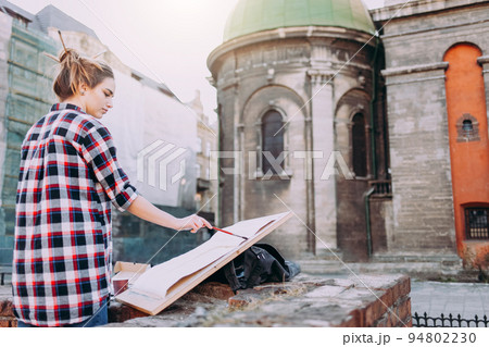 Woman is painting directly on the city street against the background of old architecture. Young painter artist in work. Woman is painting directly on the city street against the background of old architecture. Young painter artist in work. 94802230