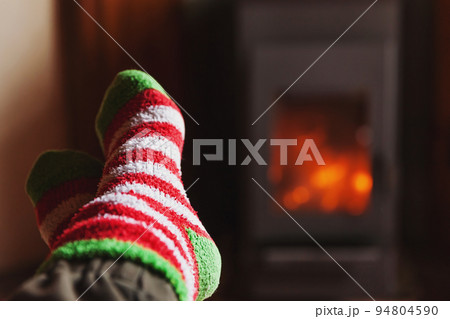 Feet legs in winter clothes wool socks at fireplace background. Woman sitting at home on winter or autumn evening relaxing and warming up. Winter and cold weather concept. Hygge Christmas eve 94804590
