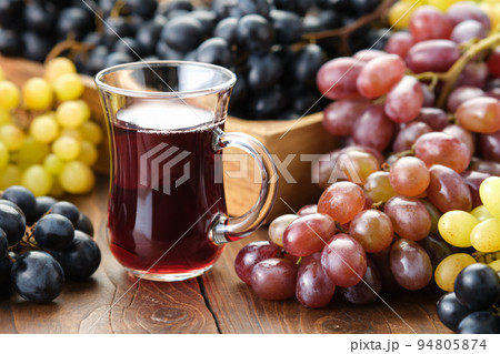 A glass cup of grape juice or wine. Black, green and purple grapes on table. 94805874