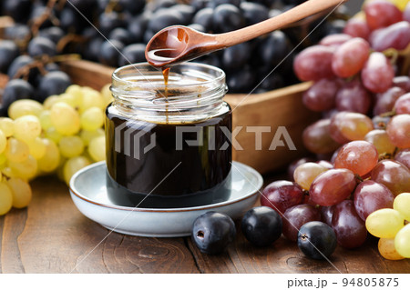 Jar of grape molasses, grape syrup. Sweetener, cough medicine. Black, green and purple grapes on kitchen table. 94805875