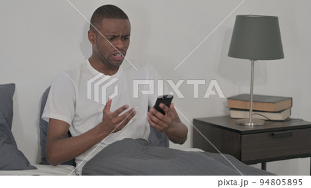 African Man Reacting to Loss on Smartphone, Sitting in Bed African Man Reacting to Loss on Smartphone, Sitting in Bed 94805895