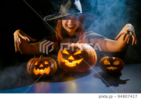 An ominous witch in a hat conjures over a jack-o-lantern. Traditional halloween characters. Mystical fog creeps over pumpkins with carved terrible faces. Woman in carnival costume. An ominous witch in a hat conjures over a jack-o-lantern. Traditional halloween characters. Mystical fog creeps over pumpkins with carved terrible faces. Woman in carnival costume. 94807427