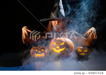 An ominous witch in a hat conjures over a jack-o-lantern. Traditional halloween characters. Mystical fog creeps over pumpkins with carved terrible faces. Woman in carnival costume. An ominous witch in a hat conjures over a jack-o-lantern. Traditional halloween characters. Mystical fog creeps over pumpkins with carved terrible faces. Woman in carnival costume. 94807428