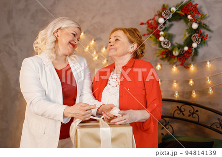 Two women celebrating Christmas together. Women sitting on a bed with gift box and posing for a photo. Red-haired old woman and her adult blonde daughter wearing red clothes. 94814139