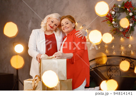 Two women celebrating Christmas together. Women sitting on a bed with gift box and posing for a photo. Red-haired old woman and her adult blonde daughter wearing red clothes. 94814141