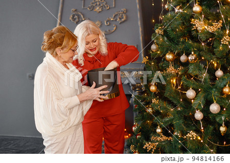 Two women celebrating Christmas together. Women standing near Christmas tree and posing for a photo. Red-haired old woman and her adult blonde daughter wearing red and white clothes. Two women celebrating Christmas together. Women standing near Christmas tree and posing for a photo. Red-haired old woman and her adult blonde daughter wearing red and white clothes. 94814166