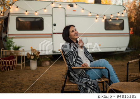Beautiful caucasian woman is sitting wrapped in a plaid and drinking a warming drink outdoors. Traveling in a motor home in autumn. Beautiful caucasian woman is sitting wrapped in a plaid and drinking a warming drink outdoors. Traveling in a motor home in autumn. 94815385