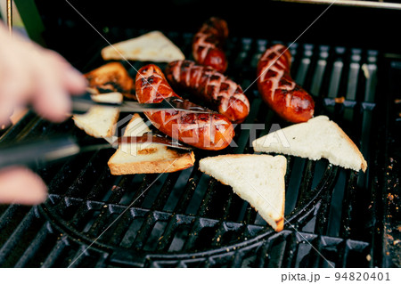 Pieces of white bread and sausages are grilling on the open fire outside. Hand of young woman grilling food, gas grill 94820401