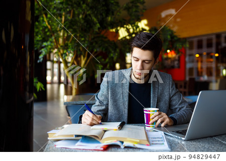 Shot of young male student sitting at table and writing on notebook. Male student studying in cafe. Shot of young male student sitting at table and writing on notebook. Male student studying in cafe. 94829447