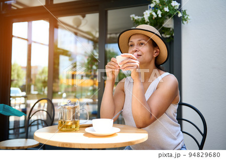 Relaxed beautiful dark-haired multiethnic woman in a straw hat and summer dress, sitting at a table outdoors in cozy cafe, enjoying a coffee break on sunny day. Lifestye. Recreation. Chill Relaxed beautiful dark-haired multiethnic woman in a straw hat and summer dress, sitting at a table outdoors in cozy cafe, enjoying a coffee break on sunny day. Lifestye. Recreation. Chill 94829680