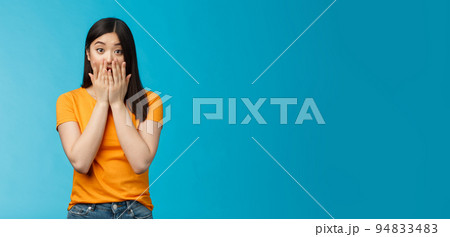 Shocked speechless stunned asian girl stare camera cover mouth palms, raise eyebrows gasping astonished, feel pitty sorry hearing bad news, stand surprised blue background wear yellow t-shirt 94833483