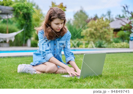 Preteen girl sitting on backyard lawn using laptop for leisure and study Preteen girl sitting on backyard lawn using laptop for leisure and study 94849745