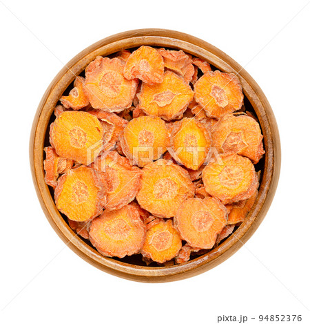 Dehydrated carrot slices in a wooden bowl. Dry disks of Daucus carota, a root vegetable with orange color. Edible taproot pieces. Close up, from above, on white background, isolated, macro food photo. 94852376