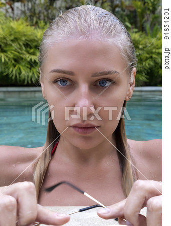 Vertical portrait of confident serious young blue-eyed blonde woman with wet hair in pool against background of blue water and plants 94854201