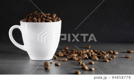 White cup with coffee beans. Gray stone background. Scattered coffee beans. 94858779