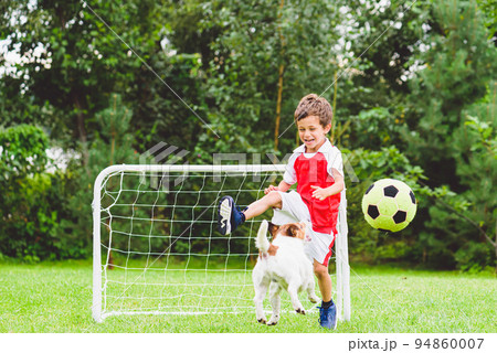 Excited child playing football (soccer) kicks ball while his dog jumping to catch it 94860007
