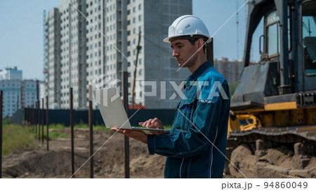 A male builder in a helmet stands against the background of a construction site with a laptop in his hands. A male builder in a helmet stands against the background of a construction site with a laptop in his hands. 94860049
