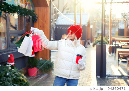 Woman walks around the city with a bunch of gift bags and drinks coffee from a disposable cup from a urban cafe. Coffee break at food kiosks after heavy shopping. choosing gifts for xmas and new year 94860471