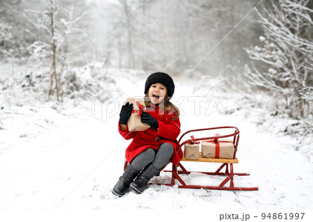A laughing five-year-old girl is sitting on a sled in a winter forest on a cold snowy day with a Christmas gift in her hands. It's time to accept and give gifts. Waiting for the New Year A laughing five-year-old girl is sitting on a sled in a winter forest on a cold snowy day with a Christmas gift in her hands. It's time to accept and give gifts. Waiting for the New Year 94861997