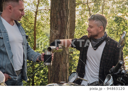 Two man friends of motorcyclists bikers on motorcycles drinking tea from a thermos on the side of the road and talking Two man friends of motorcyclists bikers on motorcycles drinking tea from a thermos on the side of the road and talking 94863120