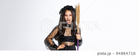 Carefree sassy asian woman enjoying halloween party, wearing witch costume and holding broom, showing tongue, standing over white background 94864710