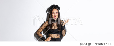 Frustrated and angry young asian woman in witch dress arguing with someone, looking pissed-off and bothered, shaking hand and looking scary at camera, standing over white background 94864711