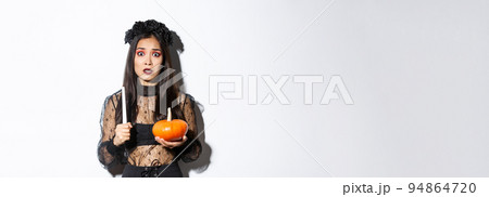 Image of asian woman panicking, being scared of witchcraft on halloween, standing in witch costume with candle and pumpkin Image of asian woman panicking, being scared of witchcraft on halloween, standing in witch costume with candle and pumpkin 94864720