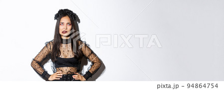Annoyed asian woman in witch costume rolling eyes and looking pressured. Irritated girl hate celebrating halloween, looking pissed-off, wearing gothic black lace dress and wreath 94864754