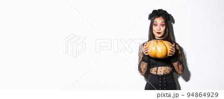 Portrait of excited pretty asian woman in halloween costume, dressed-up as witch or widow and holding pumpkin, standing over white background Portrait of excited pretty asian woman in halloween costume, dressed-up as witch or widow and holding pumpkin, standing over white background 94864929