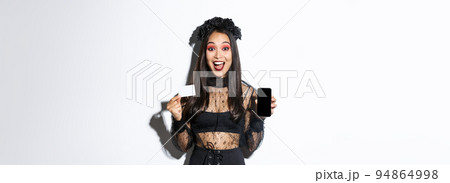 Cheerful beautiful asian woman in halloween costume, dressed as witch and showing smartphone with credit card, standing over white background Cheerful beautiful asian woman in halloween costume, dressed as witch and showing smartphone with credit card, standing over white background 94864998