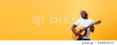 Muscular black man playing guitar, wearing jeans and white tank-top. Isolate over yellow background. 94865015