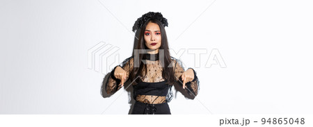 Concerned an disappointed asian woman in black lace dress and wreath smirk skeptical while pointing fingers down at something bad, complaining over white background Concerned an disappointed asian woman in black lace dress and wreath smirk skeptical while pointing fingers down at something bad, complaining over white background 94865048