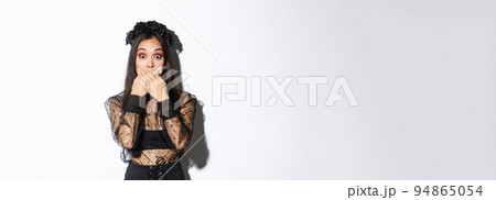 Shocked woman in witch outfit gasping and cover mouth speechless, standing over white background, looking at something surprising, wearing black lace dress and wreath, white background Shocked woman in witch outfit gasping and cover mouth speechless, standing over white background, looking at something surprising, wearing black lace dress and wreath, white background 94865054