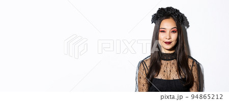 Image of smiling devious young witch looking at camera with cunning smirk, standing over white background. Woman in witch costume celebrating halloween 94865212