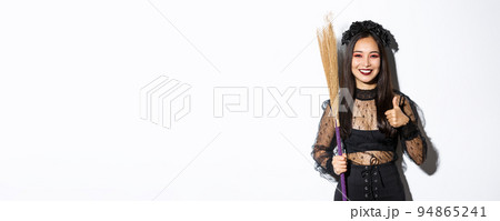 Image of smiling asian woman in witch costume with broom, showing thumbs-up in approval, standing over white background 94865241
