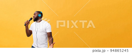 Portrait of cheerful positive chic handsome african man holding microphone and having headphones on head listening music singing song enjoying weekend vacation isolated on yellow background 94865245