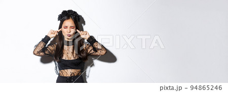 Annoyed asian woman in witch costume, close eyes and shut ears with fingers, unwilling to listen, bothered by loud noise, standing in gothic dress and wreath over white background 94865246
