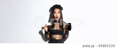 Amazed asian girl in witch costume showing credit card with mobile phone screen, open mouth surprised, celebrating halloween, white background 94865320