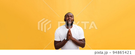 Studio portrait of young African American man in white shirt, holding hands in prayer, looking at the camera with thoughtful skeptical expression on his face, suspecting of something. Body language. 94865421