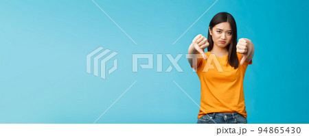 Upset gloomy asian girl with dark haircut frowning sulking sad, disappointed, show thumbs-down grimacing, give negative opinion, judging bad uninteresting movie, stand blue background 94865430
