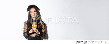 Image of funny carefree girl celebrating halloween in witch costume, making silly faces and holding treats, standing over white background 94865495