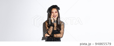 Image of happy good-looking asian woman in gothic dress and black wreath, laughing at halloween party, standing over white background Image of happy good-looking asian woman in gothic dress and black wreath, laughing at halloween party, standing over white background 94865579