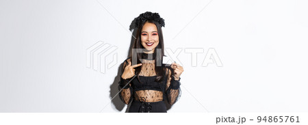 Image of confident smiling asian woman in witch gothic costume pointing finger at credit card, standing white background 94865761