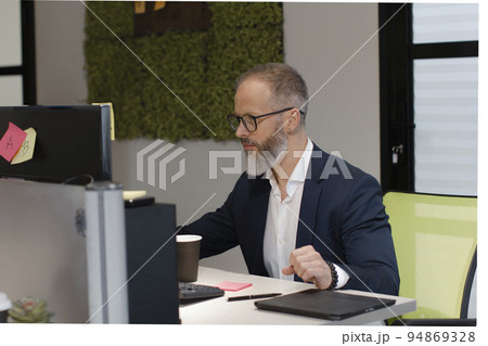 A European adult man with a beard works at a computer in the office of company A European adult man with a beard works at a computer in the office of company 94869328