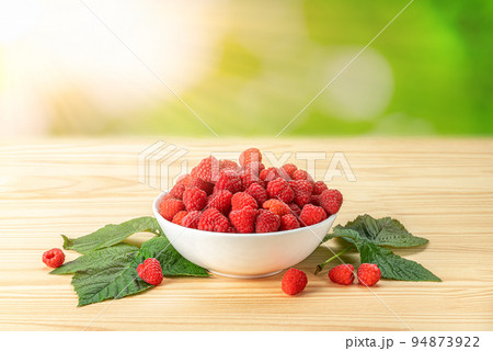 Raspberry on white plate on wooden table at sunny day. Plate with ripe red raspberries stands on wooden table against blurred background of greenery. Fresh organic ripe raspberry. Raspberry on white plate on wooden table at sunny day. Plate with ripe red raspberries stands on wooden table against blurred background of greenery. Fresh organic ripe raspberry. 94873922