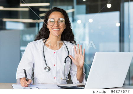 Portrait of a Latin American female pediatrician doctor. Sits in the office and conducts online consultations with patients. He looks into the camera, waves his hand, writes a medical history, smiles. 94877677