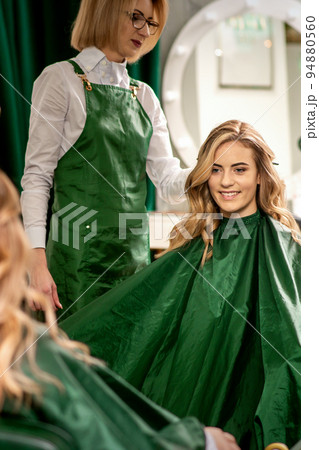Hairdresser combing long hair of young caucasian woman looking and smiling in the mirror in beauty salon Hairdresser combing long hair of young caucasian woman looking and smiling in the mirror in beauty salon 94880560